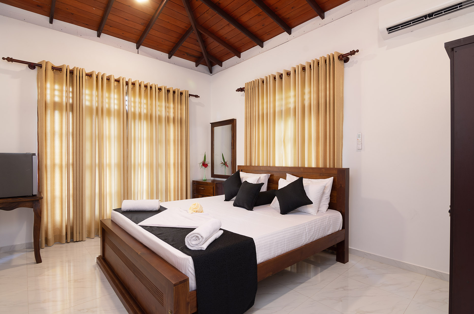 Elegant bedroom with a large bed featuring white bedding and black accent pillows, golden curtains over windows, and vaulted ceiling with exposed wooden beams. Includes modern amenities like a mini fridge and full-length mirror, creating a luxurious and inviting atmosphere