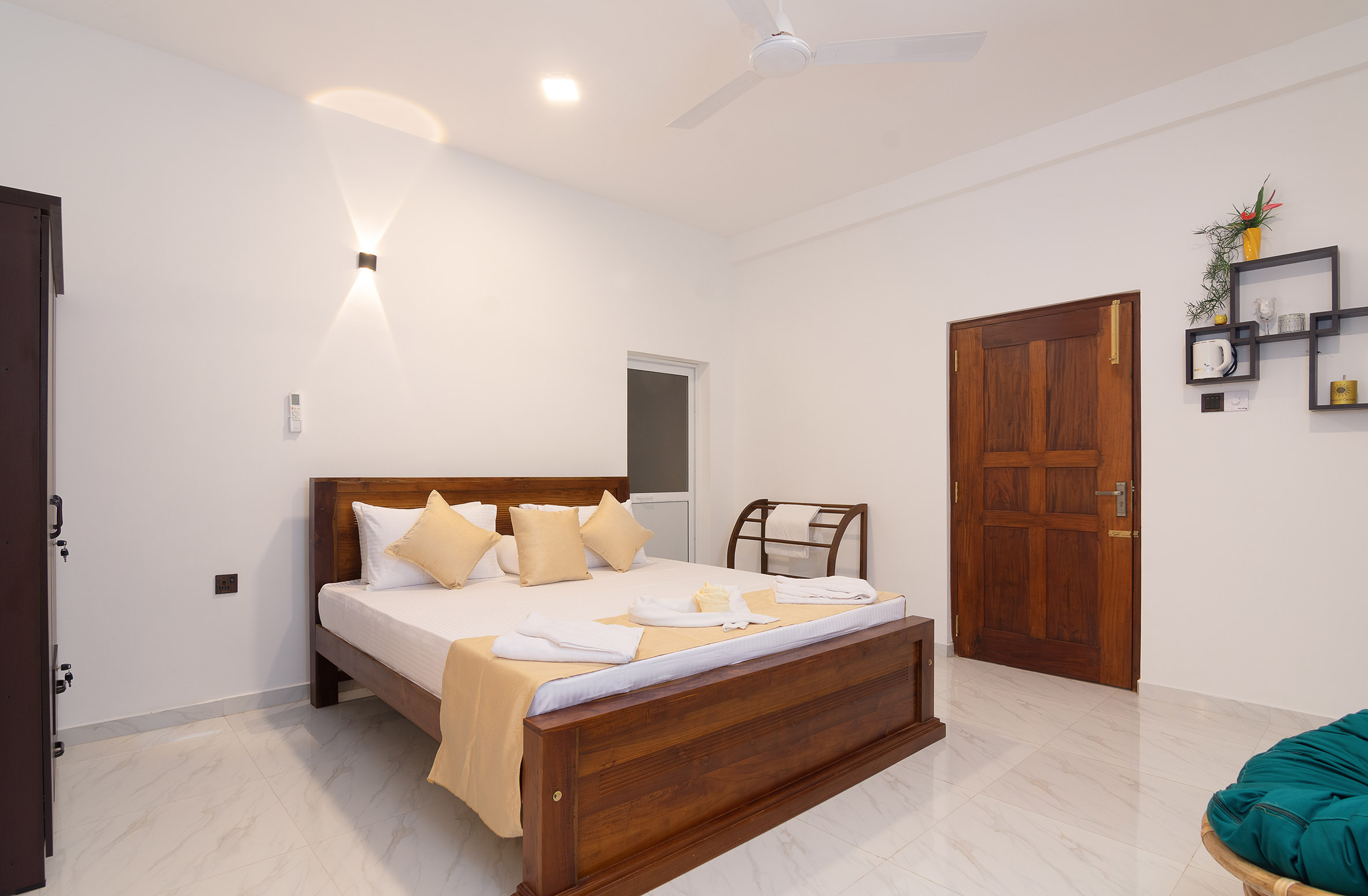 A well-lit, spacious bedroom with white walls and a large window. The room features a double bed with a wooden headboard, white bedding, and four yellow pillows. Two white towels are placed at the foot of the bed. Above the bed is a ceiling fan, and to the right is a small wooden chair with clothes draped over it. On the left wall, there’s a flat-screen TV mounted above a dark wood console table. The right wall has an open door leading out of the bedroom and a small shelf with decorative items including plants and framed pictures. The floor is tiled in light beige, complementing the room’s neutral color palette