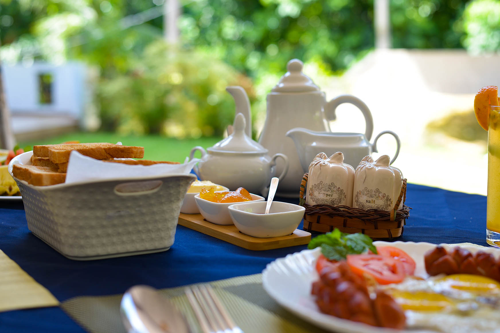 Indulge in a delicious breakfast at Tranquil Escape villa in Hikkaduwa, Sri Lanka. Enjoy freshly baked toast, eggs, sausages, and refreshing beverages served on the outdoor terrace with stunning garden views.