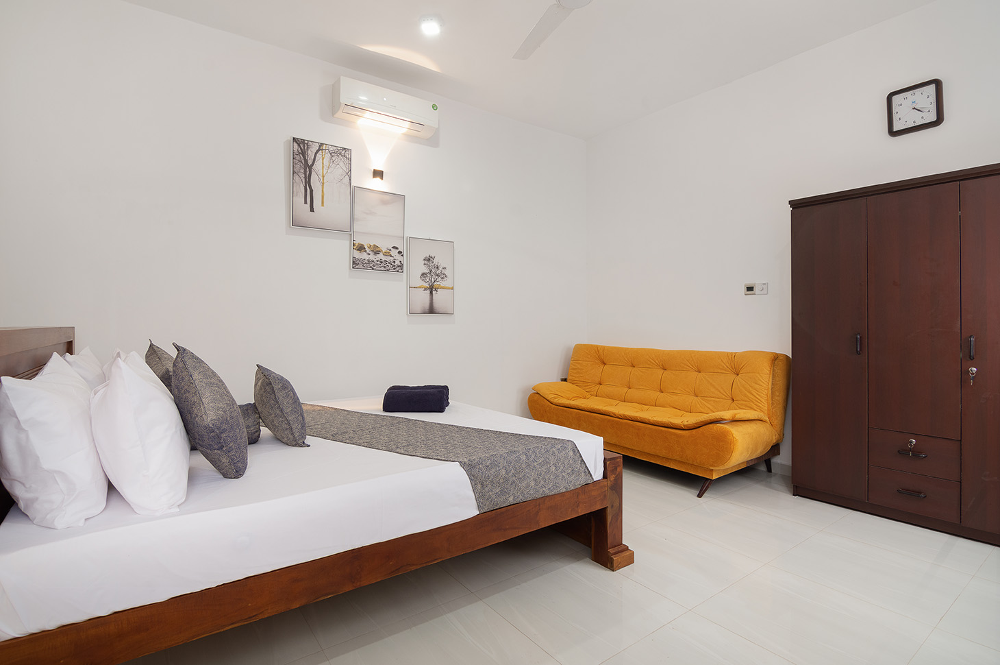 Spacious and modern villa room featuring a king-sized bed, a cozy mustard-colored sofa, and elegant minimalist decor, offering a comfortable and relaxing stay