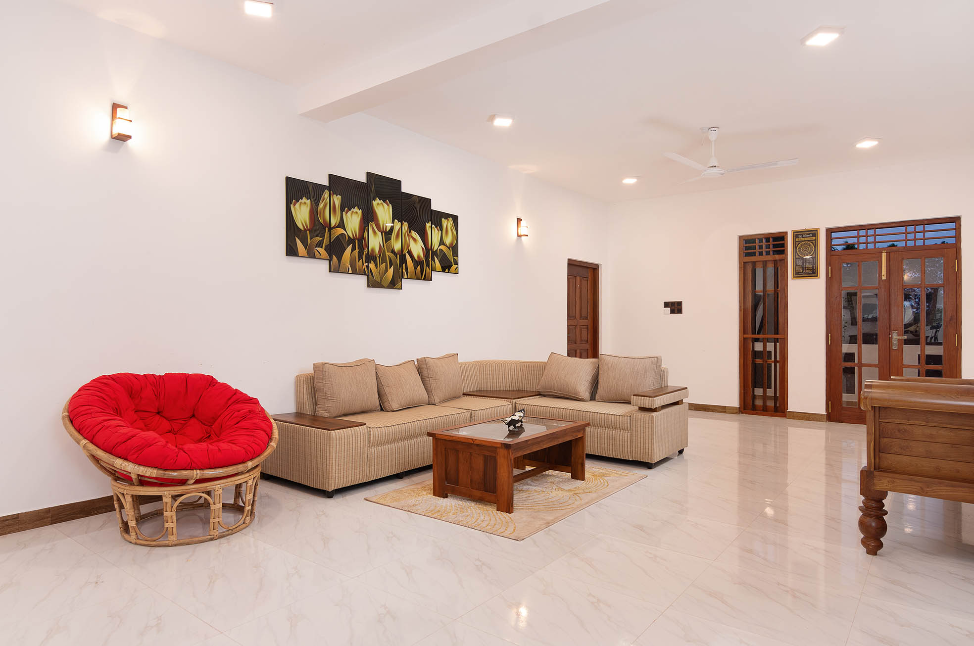 Tranquil Escape Hikkaduwa: Spacious living room with beige L-shaped sofa, vibrant red papasan chair, wooden coffee table, and floral wall art. Bright, airy space with polished marble floors, recessed lighting, and elegant wooden doors leading to outdoor areas