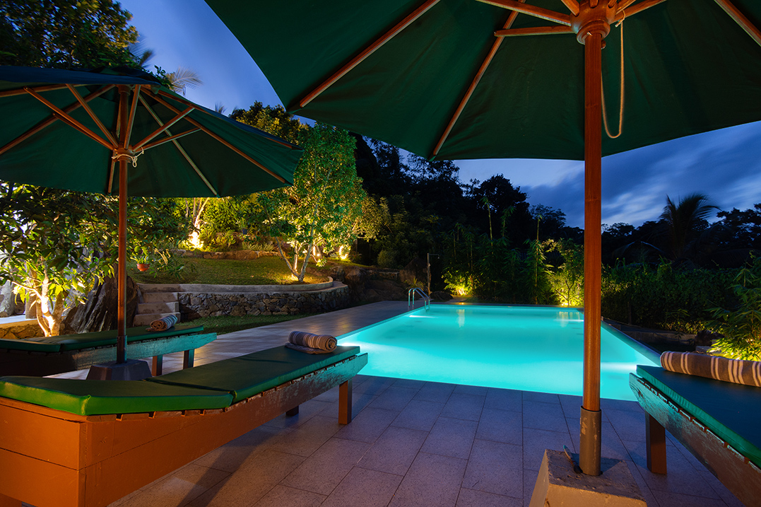 A serene outdoor pool area at dusk, featuring illuminated pool water, green umbrellas, lounge chairs with green cushions, and surrounding trees with soft lighting.