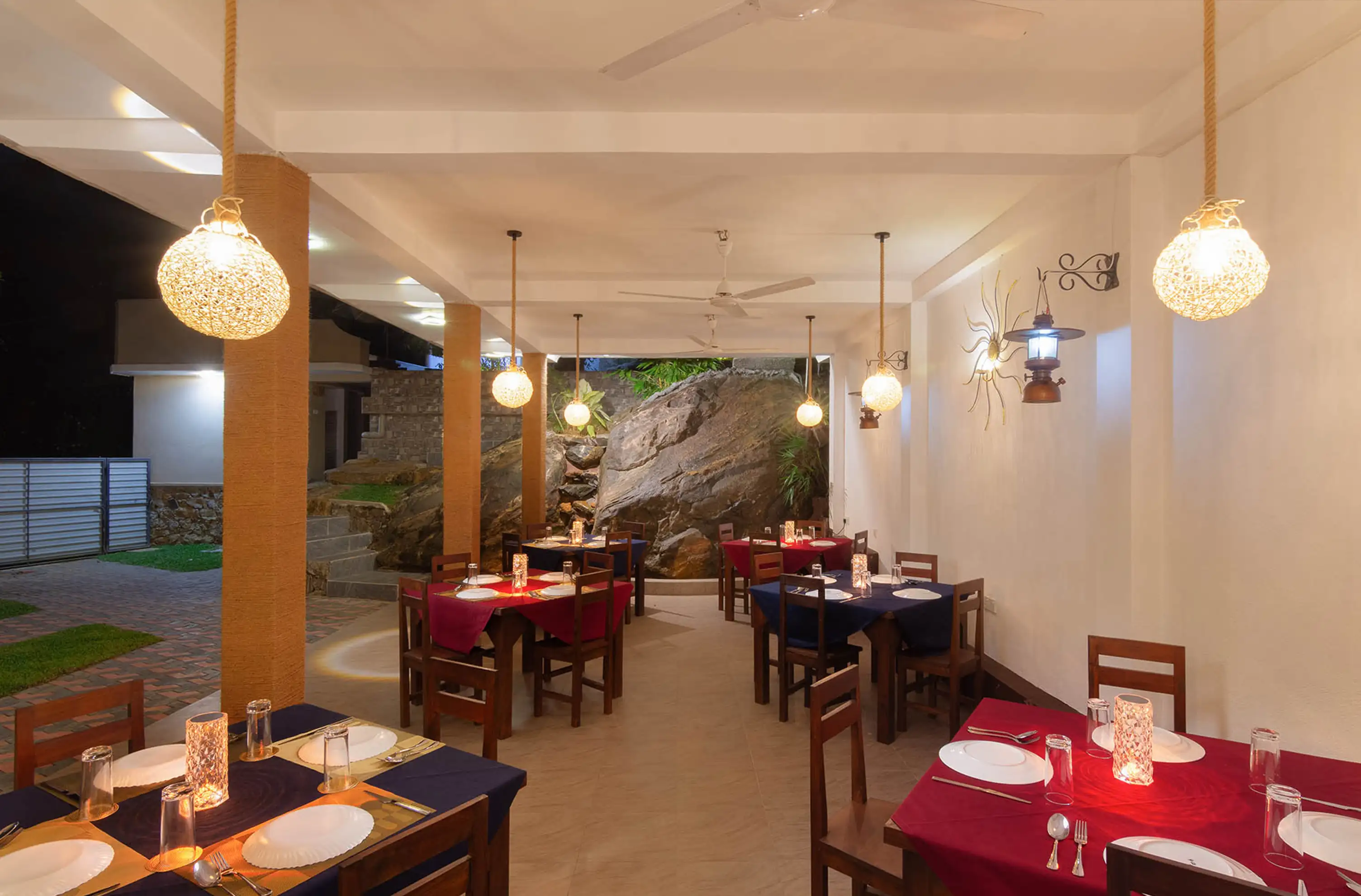 Indoor dining area featuring contemporary design with multiple tables set with white plates, silverware, and burgundy tablecloths. Spherical pendant lights illuminate the space, which includes light-colored tile flooring and a large rock formation wall, creating a stylish and comfortable dining environment