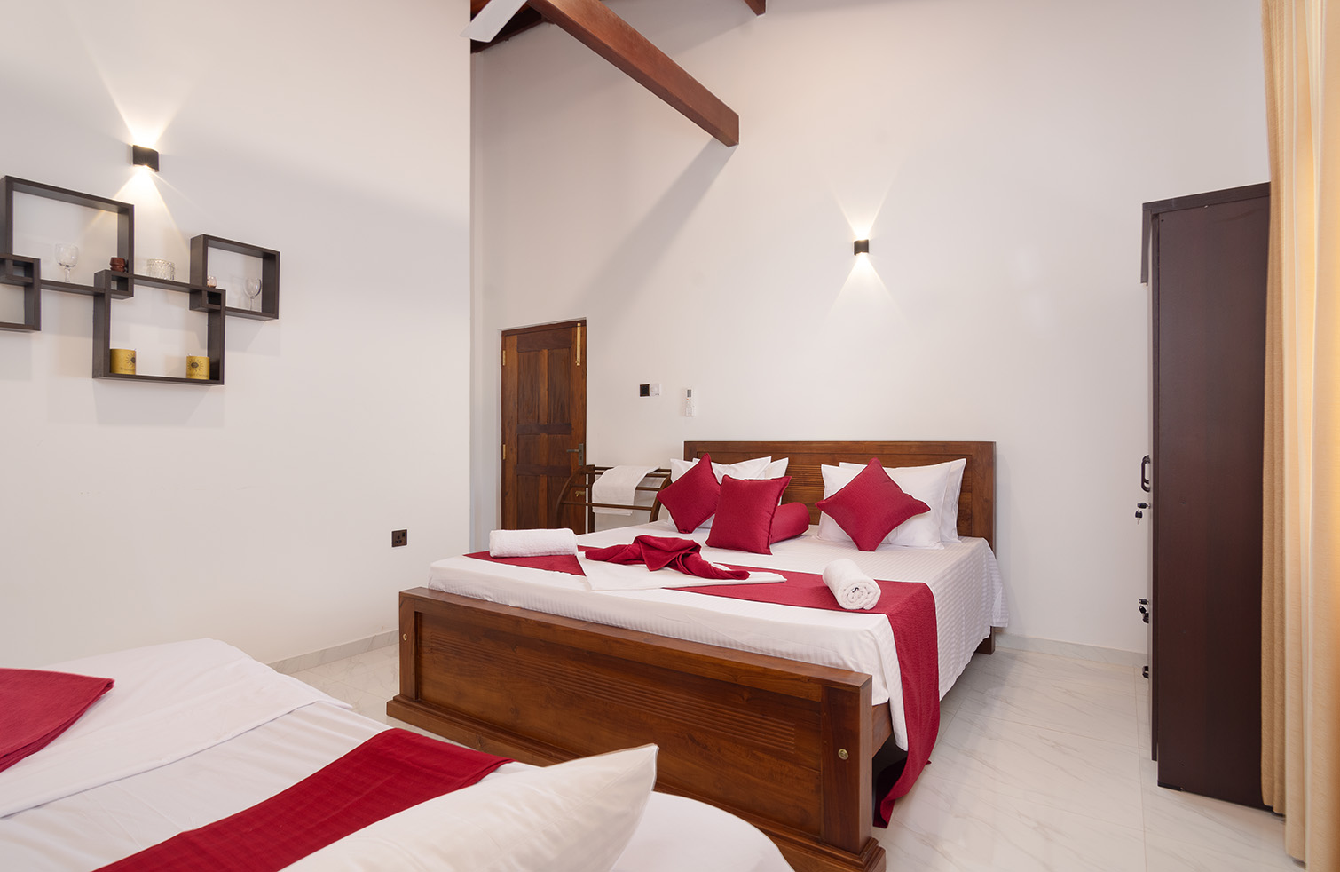 Neatly arranged bedroom with a large bed featuring a white duvet and red accent pillows, two smaller beds to the side, and wall-mounted lights. The room has white walls, a sloped ceiling with exposed wooden beams, and a dark wooden door, creating a cozy and inviting atmosphere.